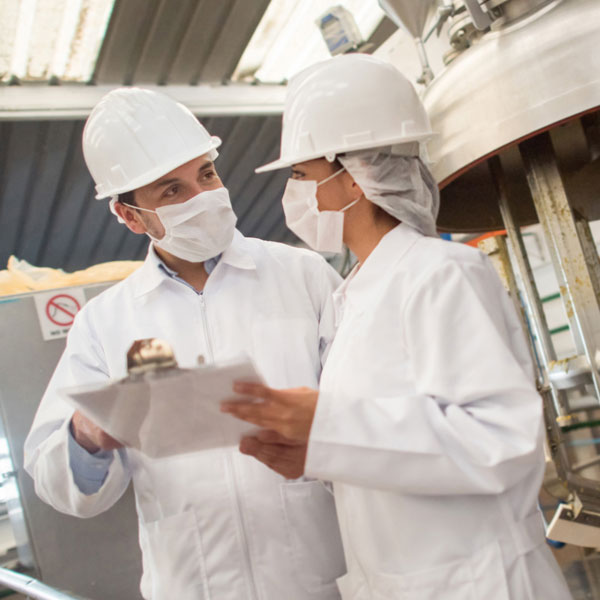 Two inspectors are in a food manufacturing facility. They are in protective equipment and holding a clipboard.
