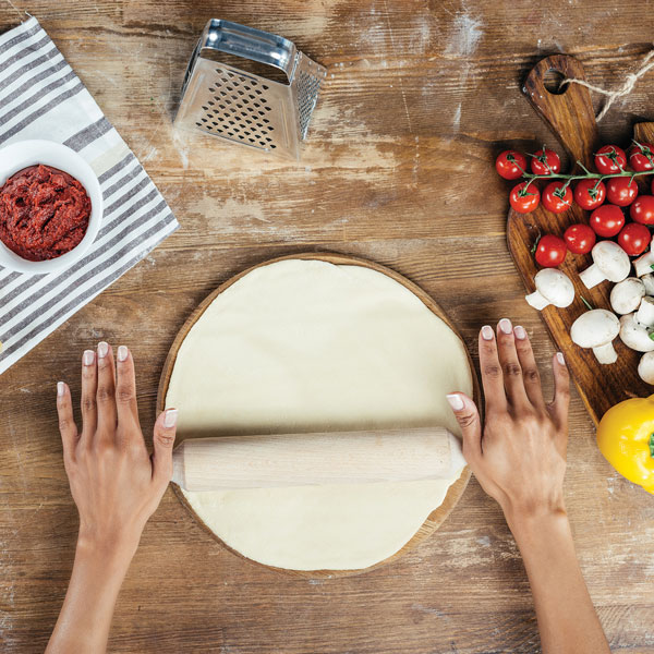Two hands are rolling out pizza dough. Around the dough there is cheese, tomato sauce, and ingredients needed to make a pizza.