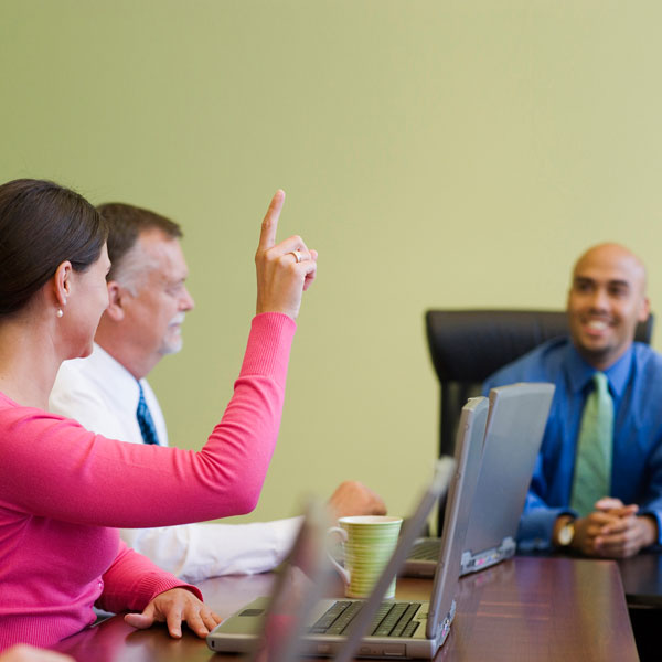 Trois personnes assises à une table de réunion. Une femme lève la main pour poser une question.
